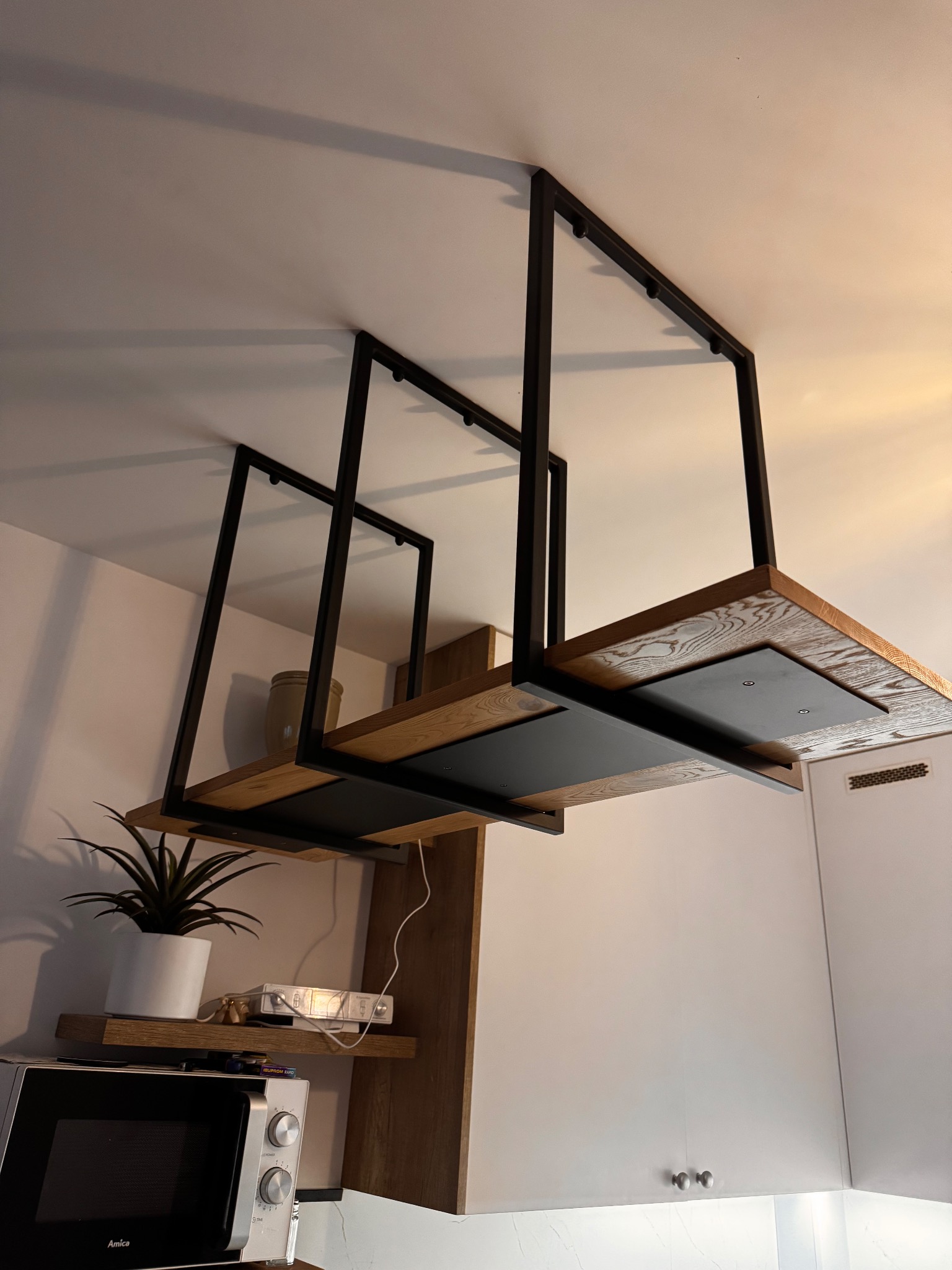 Nowoczesna, podwieszana półka z drewnianymi blatami i czarną, metalową konstrukcją, zamontowana w kuchni. Widoczny fragment zabudowy kuchennej i sprzęt AGD.