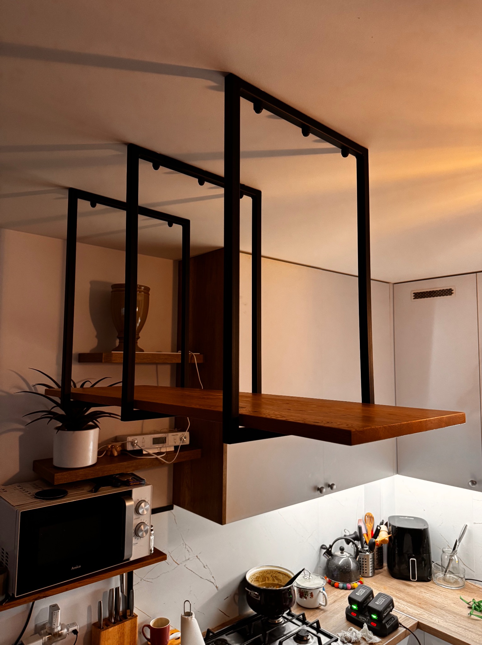 Nowoczesna, metalowa konstrukcja podwieszana w kuchni, z drewnianymi półkami. Minimalistyczny design, czarne profile, ciepłe oświetlenie. Widok na kuchenny blat i sprzęty.