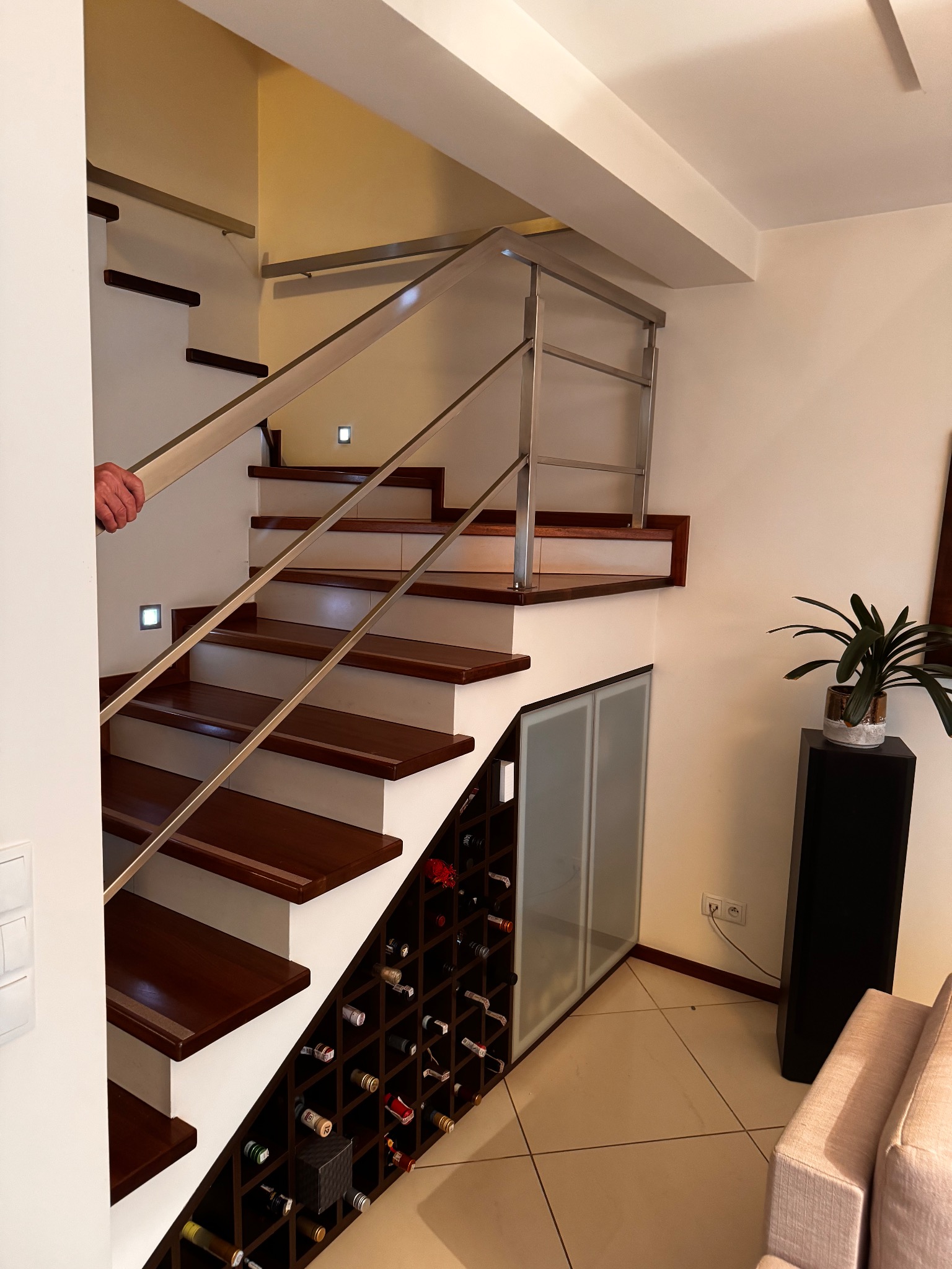 Nowoczesne schody drewniane z metalową balustradą i wbudowaną szafką na wino, oświetlone punktowo, w jasnym wnętrzu domu.