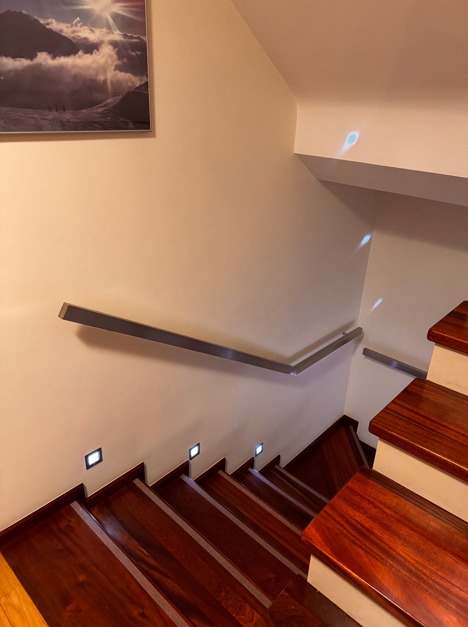 Drewniane schody z metalową poręczą i oświetleniem punktowym w ścianie. Na ścianie wisi obraz z motywem górskim. Ujęcie z góry, podkreślające detale wykończenia.
