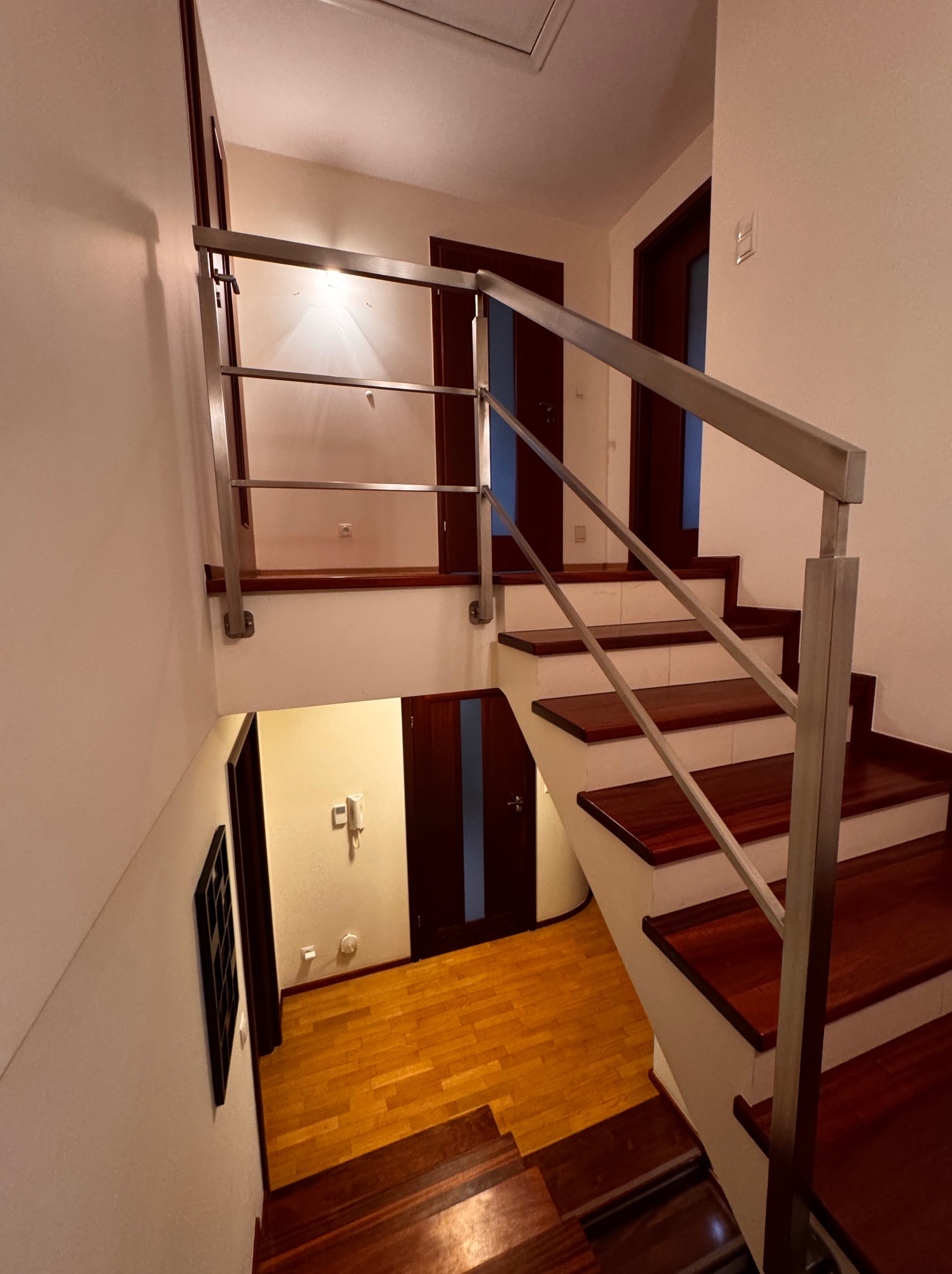 Wnętrze domu z drewnianymi schodami i nowoczesną, metalową poręczą. Widok z góry na dół, ukazujący detale wykończenia i oświetlenie.