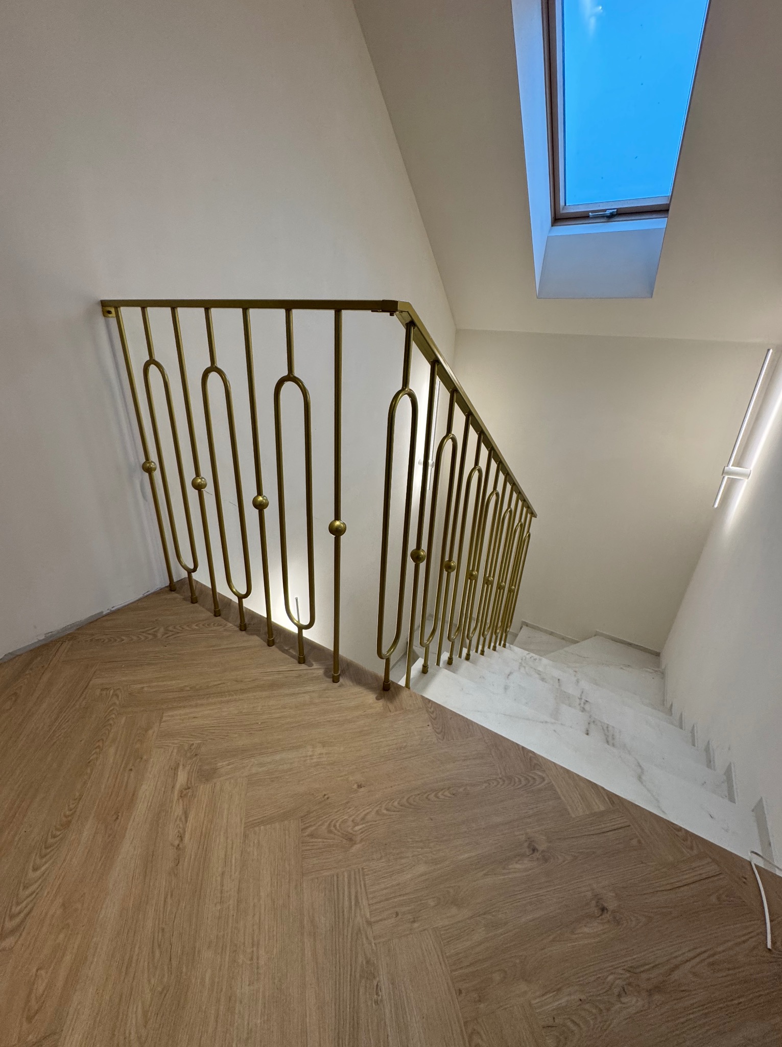 Złota, metalowa barierka schodowa z dekoracyjnymi elementami na tle białych ścian, drewnianej podłogi i marmurowych schodów. Widok z góry, oświetlenie naturalne.