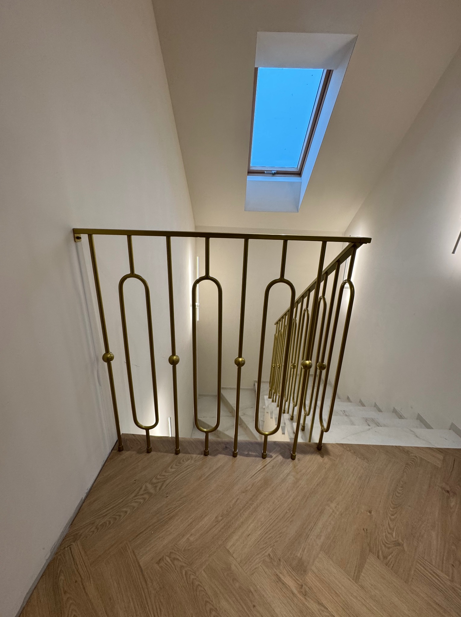 Złota, dekoracyjna poręcz schodowa z motywem owalnym, widziana z góry, na tle drewnianej podłogi w jodełkę i marmurowych schodów. W tle okno dachowe.