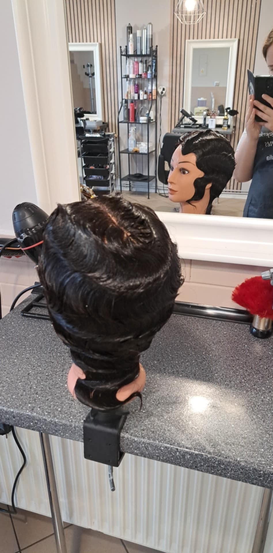 Manekin fryzjerski z ułożoną fryzurą w stylu retro, widoczny w lustrze. Na stole suszarka i akcesoria. Precyzyjne fale na ciemnych włosach.