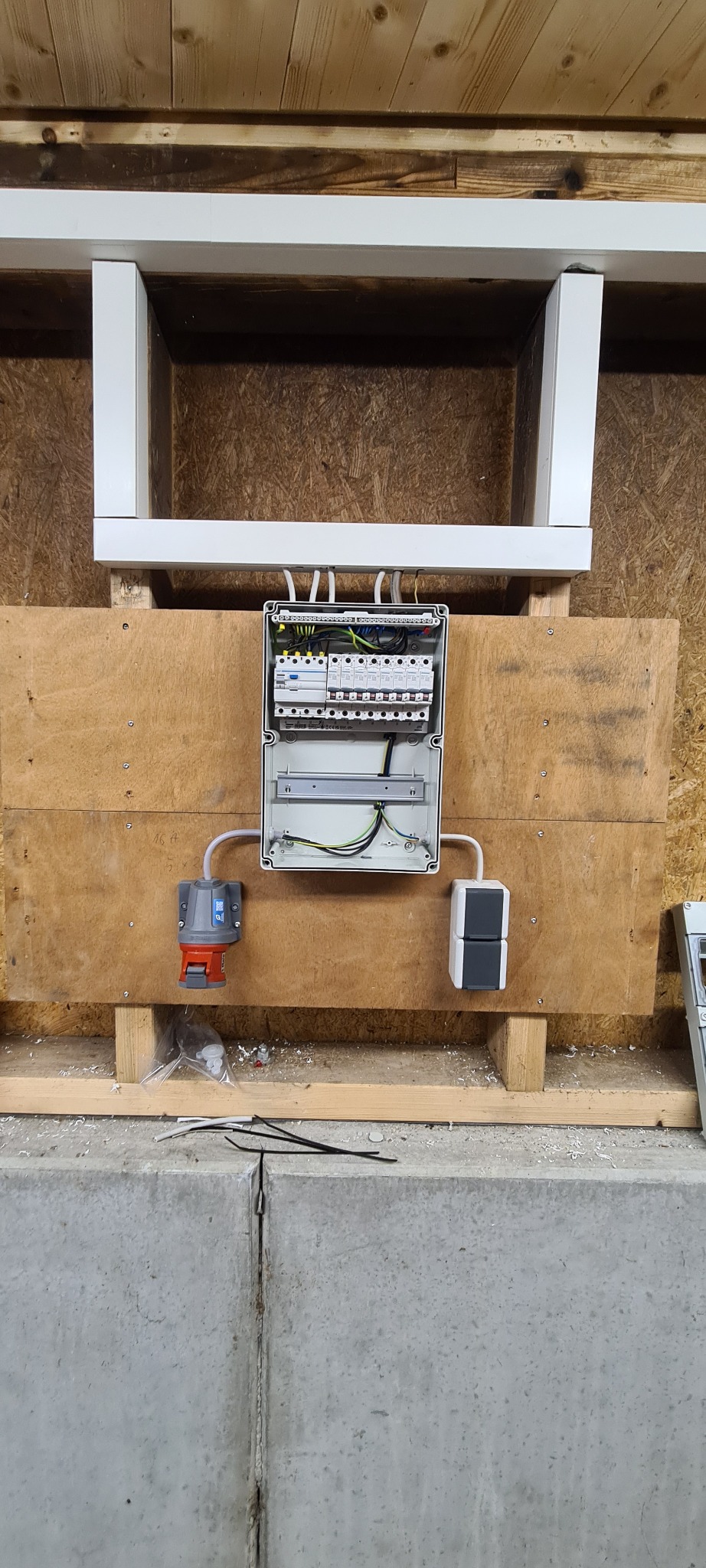 Otwarta skrzynka elektryczna z widocznymi bezpiecznikami i przewodami, zamontowana na drewnianej płycie. Dwa gniazda po bokach, przewody i elementy montażowe na podłodze.