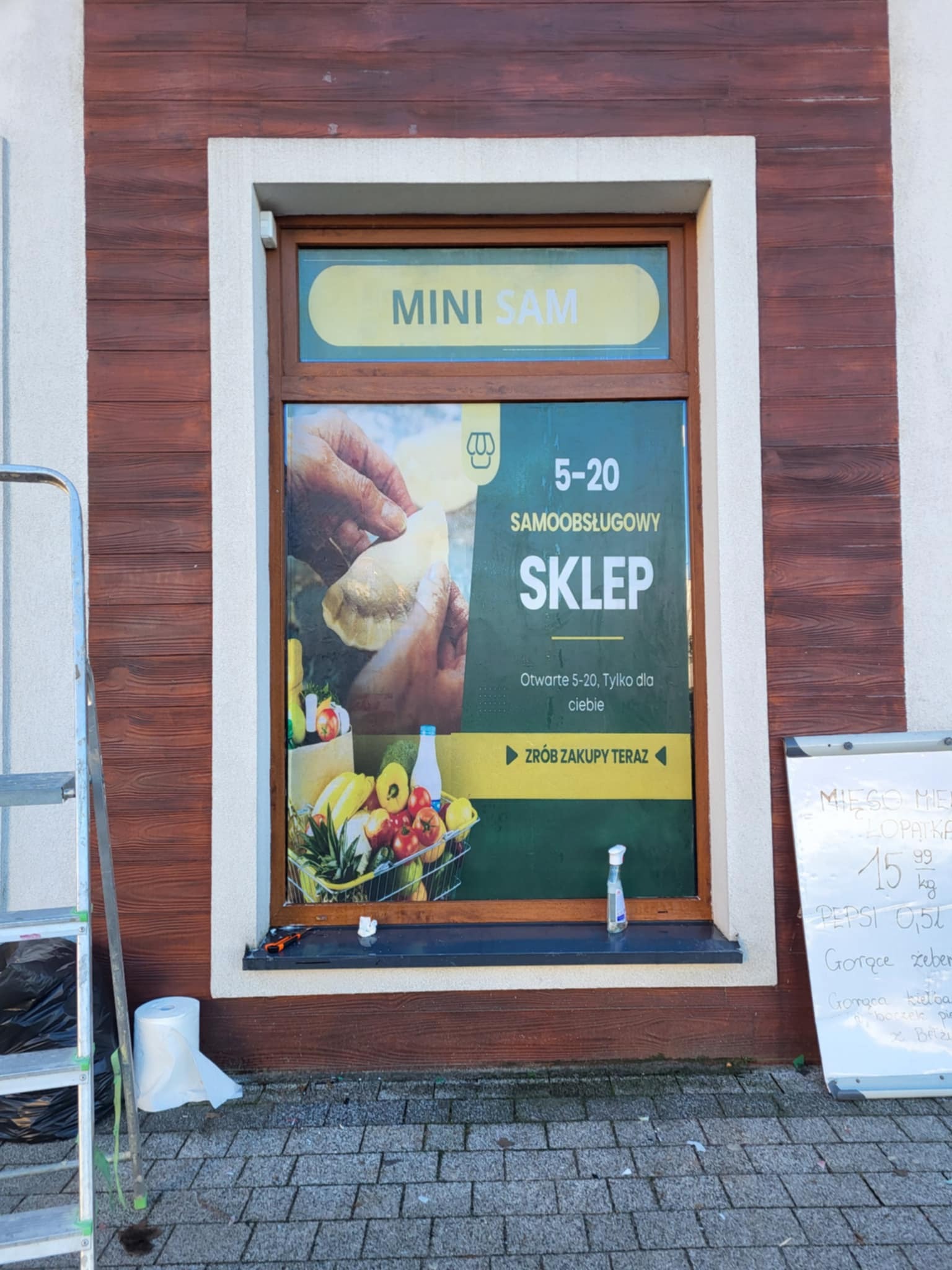 Okno sklepu 'Mini Sam' z naklejoną reklamą, widoczna drabina i tablica z ofertą. Ujęcie z zewnątrz, w tle brązowa elewacja imitująca drewno.