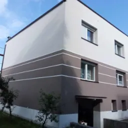 Dwukondygnacyjny dom z elewacją w odcieniach brązu i beżu, z białymi pasami. Widoczne okna z roletami, fragment rusztowania i zieleń wokół budynku.