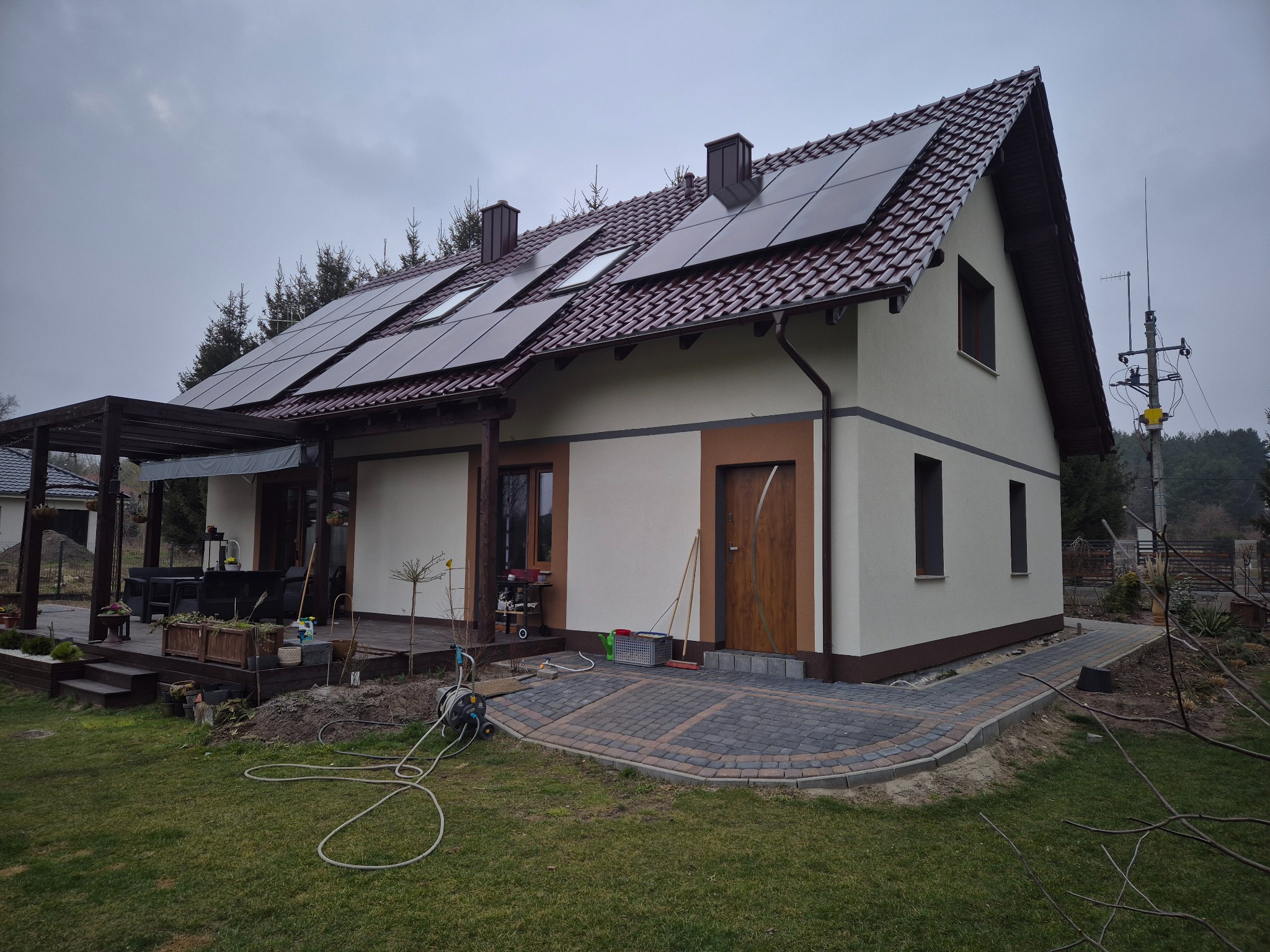 Jasny dom jednorodzinny z panelami słonecznymi na dachu i nową elewacją w odcieniach beżu i brązu. Widoczne wykończenie z kostki brukowej i elementy ogrodu.