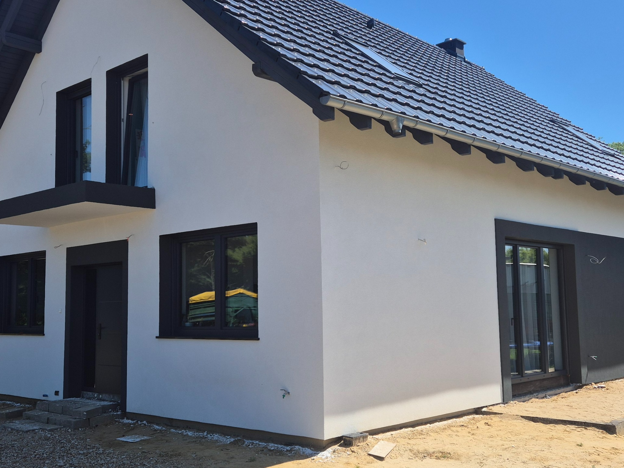 Elewacja domu jednorodzinnego z białym tynkiem i czarnymi ramami okiennymi, widok z boku na tle błękitnego nieba. Nowoczesny design z elementami wykończeniowymi.