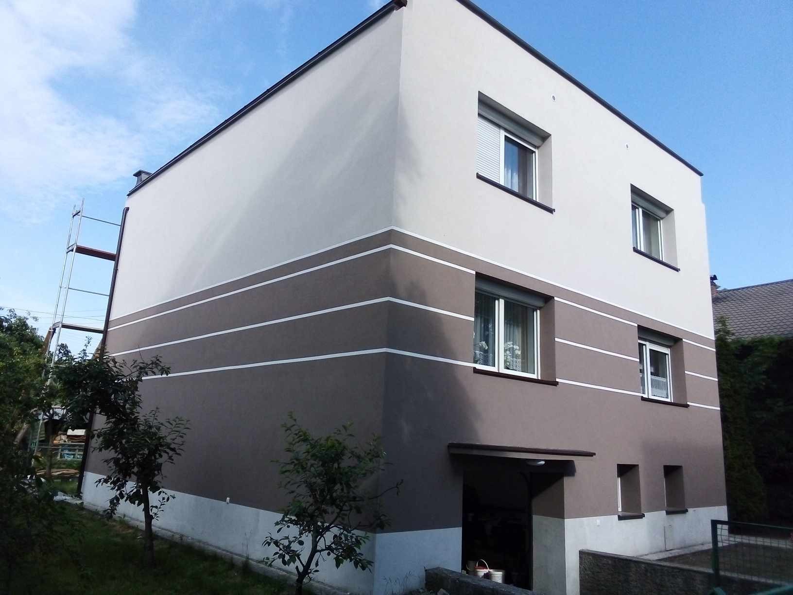 Dwukondygnacyjny dom z elewacją w odcieniach brązu i beżu, z białymi pasami. Widoczne okna z roletami, fragment rusztowania i zieleń wokół budynku.