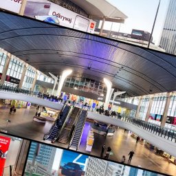 CITYPHONIC MEDIAHUB SPÓŁKA Z OGRANICZONĄ ODPOWIEDZIALNOŚCIĄ - Wnętrze hali dworcowej z reklamami na ścianach i billboardach. Widoczne ruchome schody i osoby. Panorama miasta w tle. Kolaż.
