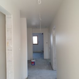 Usługi Budowlano-Remontowe Paweł Wojciechowski - Wnętrze w trakcie malowania: świeżo pomalowane ściany w jasnym odcieniu, widoczny kubeł z farbą, surowy beton na podłodze i wystające kable elektryczne.