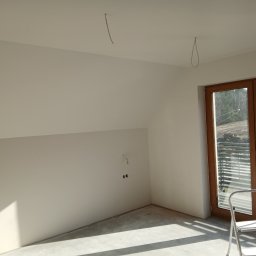 Usługi Budowlano-Remontowe Paweł Wojciechowski - Poddasze po szpachlowaniu i malowaniu na biało, widoczne wystające kable i drabina oparta o okno balkonowe. Ściany gładkie, przygotowane do dalszych prac wykończeniowych.