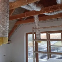 Montaż izolowanych przewod&oacute;w wentylacyjnych na poddaszu domu jednorodzinnego z drewnianą konstrukcją dachu, widoczne rusztowanie.