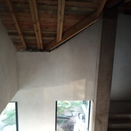 Usługi Budowlano-Remontowe Paweł Wojciechowski - Surowe wnętrze z drewnianą konstrukcją dachu i otynkowanymi ścianami, widoczne okno z widokiem na zewnątrz. Etap prac wykończeniowych w domu.