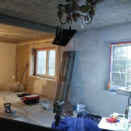 Usługi Budowlano-Remontowe Paweł Wojciechowski - Wnętrze w trakcie remontu: surowy strop, ściany po szpachlowaniu, okna z folią ochronną, drabina oparta o ścianę, narzędzia i materiały budowlane.