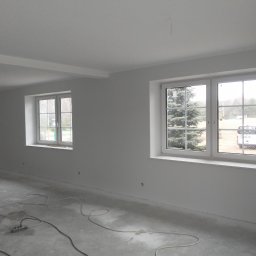 Usługi Budowlano-Remontowe Paweł Wojciechowski - Białe, świeżo pomalowane wnętrze z dwoma oknami i szarym betonowym podłożem. Widoczne kable i listwy przypodłogowe. Minimalistyczny styl, surowy stan wykończenia.