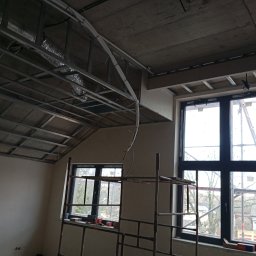 Usługi Budowlano-Remontowe Paweł Wojciechowski - Stelaż sufitu podwieszanego z widoczną instalacją wentylacyjną i elektryczną w surowym wnętrzu. Dwa okna z widokiem na zewnątrz. Metalowe rusztowanie w pomieszczeniu.