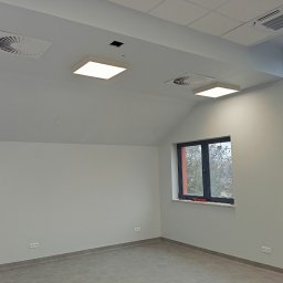 Usługi Budowlano-Remontowe Paweł Wojciechowski - Jasne, świeżo pomalowane biuro z dwoma oknami, wentylacją sufitową i nowoczesnym oświetleniem. Widoczne gładkie ściany i szara podłoga.