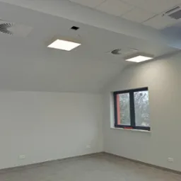 Jasne, świeżo pomalowane biuro z dwoma oknami, wentylacją sufitową i nowoczesnym oświetleniem. Widoczne gładkie ściany i szara podłoga.