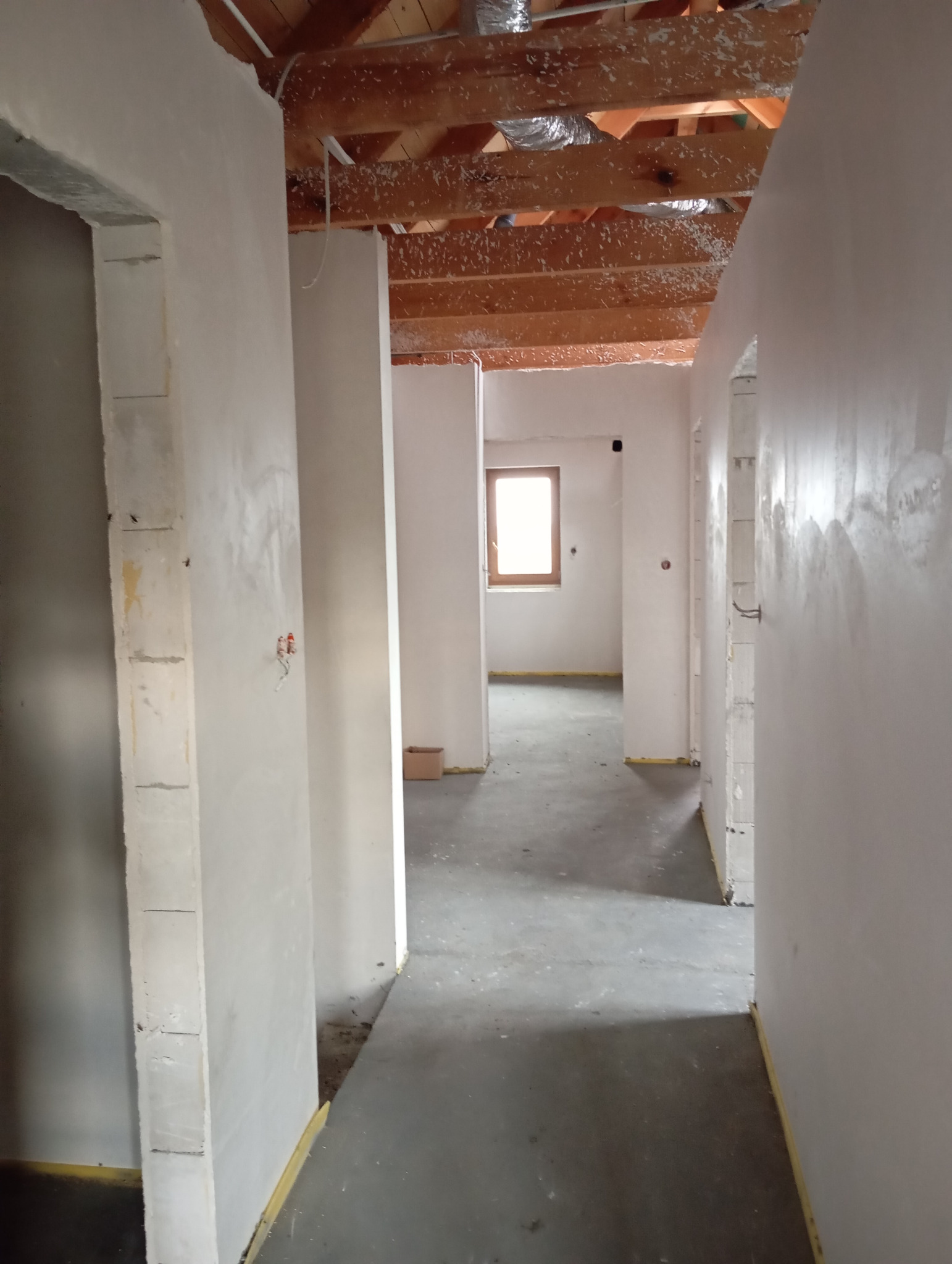 Wnętrze w trakcie remontu: surowe ściany po szpachlowaniu, widoczne drewniane belki stropowe z resztkami izolacji. Okno w głębi korytarza. Stan surowy.