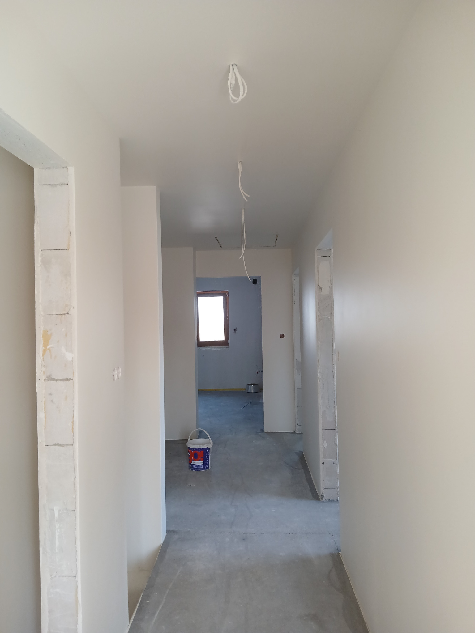 Wnętrze w trakcie malowania: świeżo pomalowane ściany w jasnym odcieniu, widoczny kubeł z farbą, surowy beton na podłodze i wystające kable elektryczne.