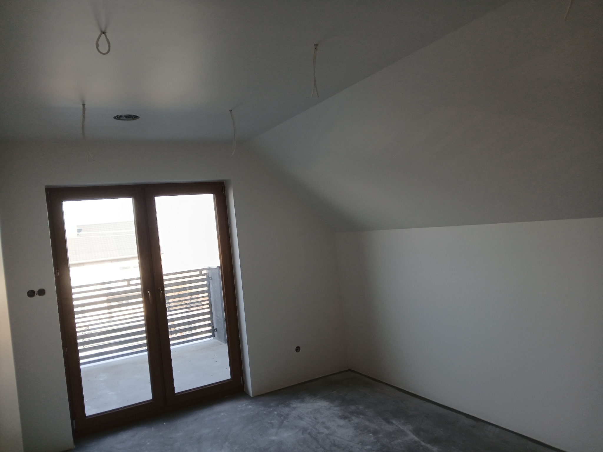 Poddasze po szpachlowaniu i malowaniu ścian na biało, z widocznymi przewodami oświetleniowymi i wyjściem na balkon z drewnianą ramą okienną.