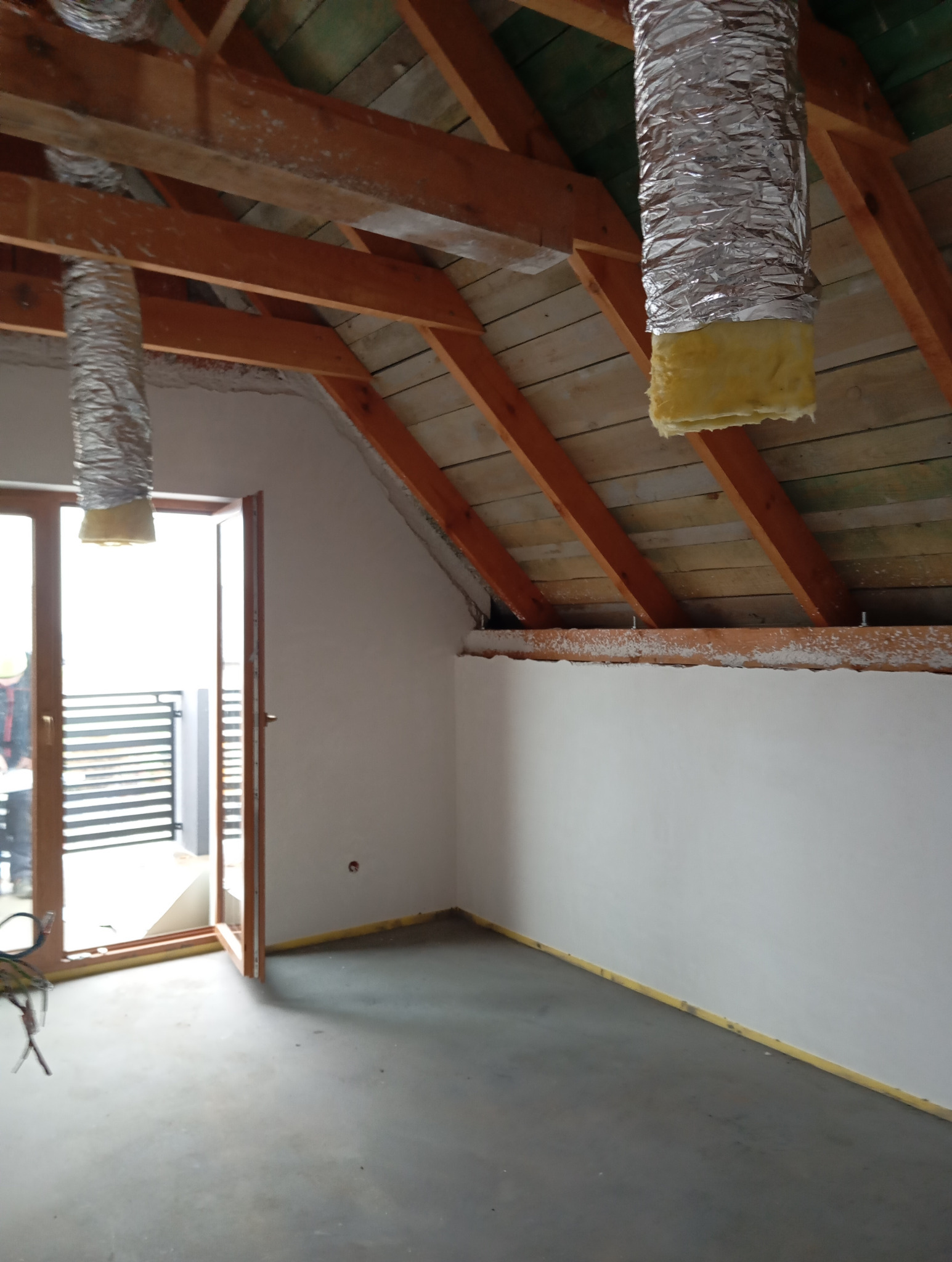 Poddasze w trakcie adaptacji: surowe ściany, drewniana konstrukcja dachu, widoczne przewody wentylacyjne z izolacją, otwarte drzwi balkonowe w tle.