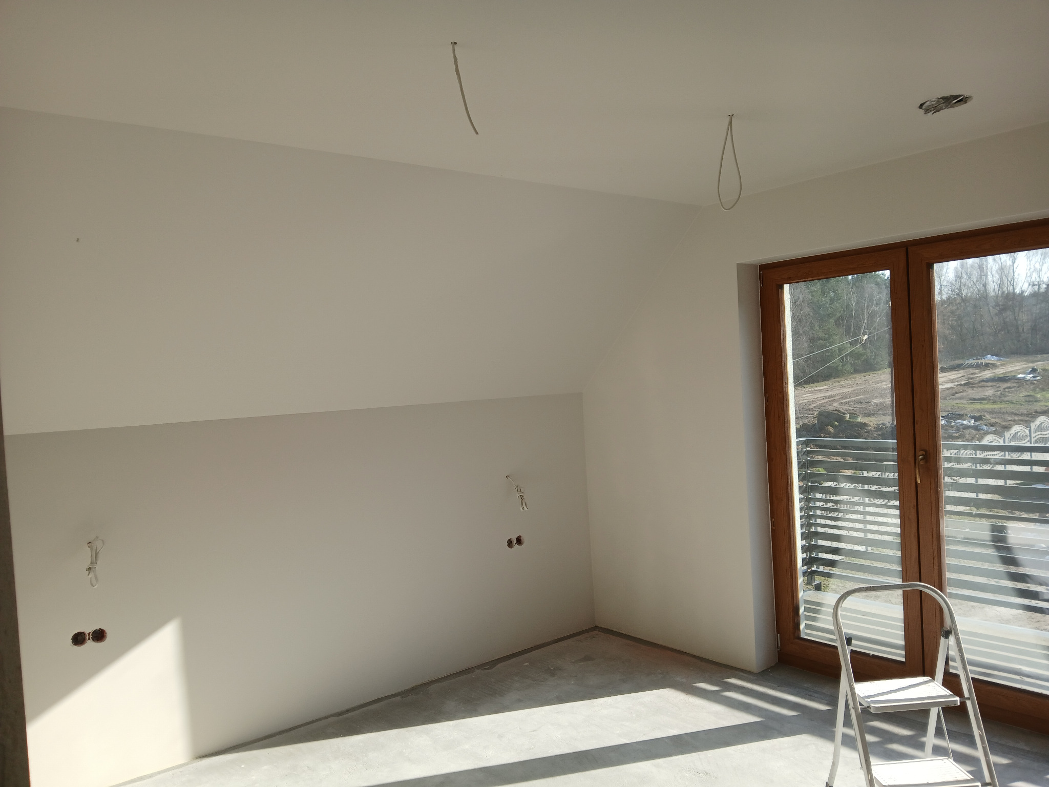 Poddasze po szpachlowaniu i malowaniu na biało, widoczne wystające kable i drabina oparta o okno balkonowe. Ściany gładkie, przygotowane do dalszych prac wykończeniowych.