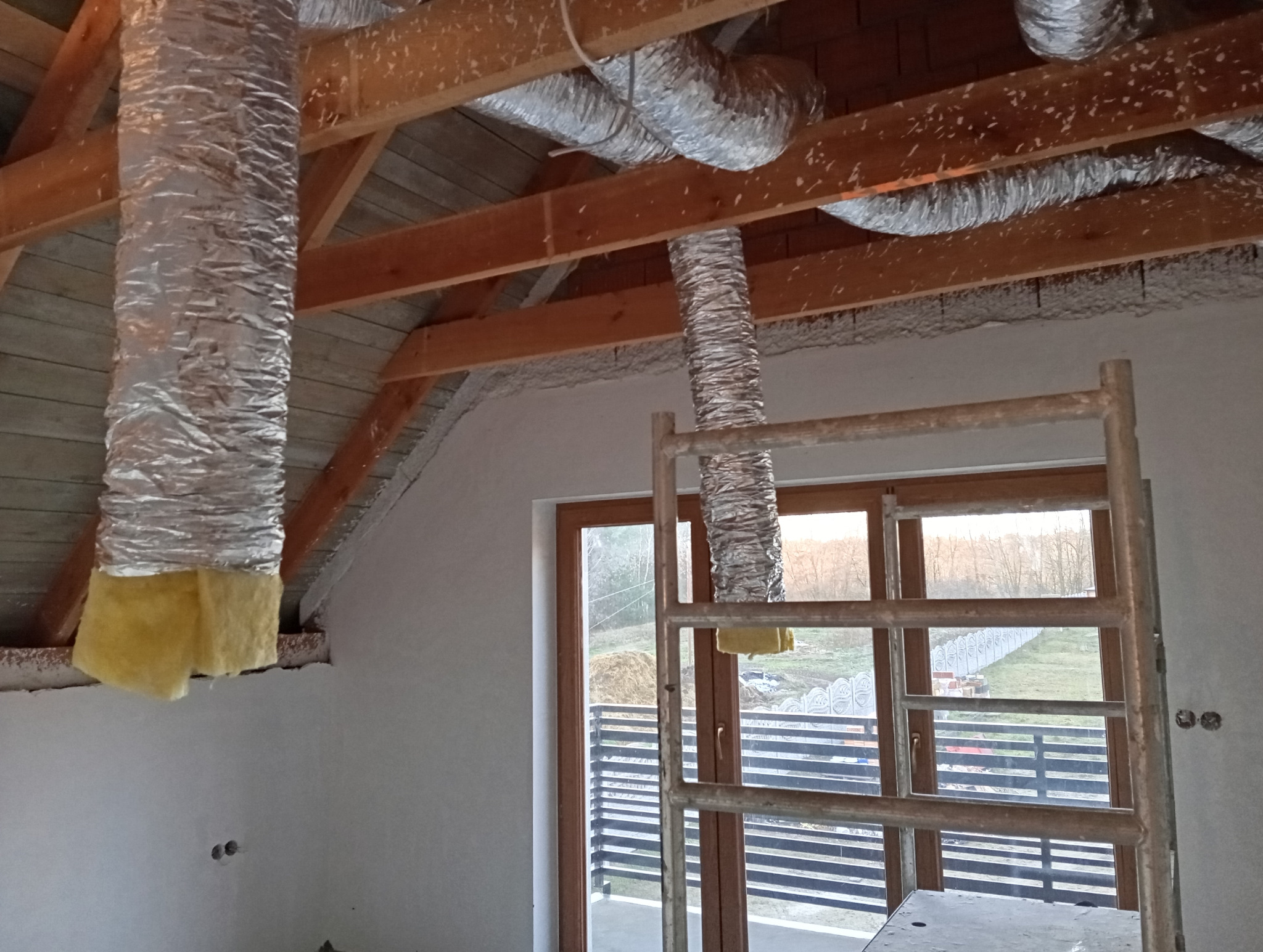 Montaż izolowanych przewodów wentylacyjnych na poddaszu domu jednorodzinnego z drewnianą konstrukcją dachu, widoczne rusztowanie.