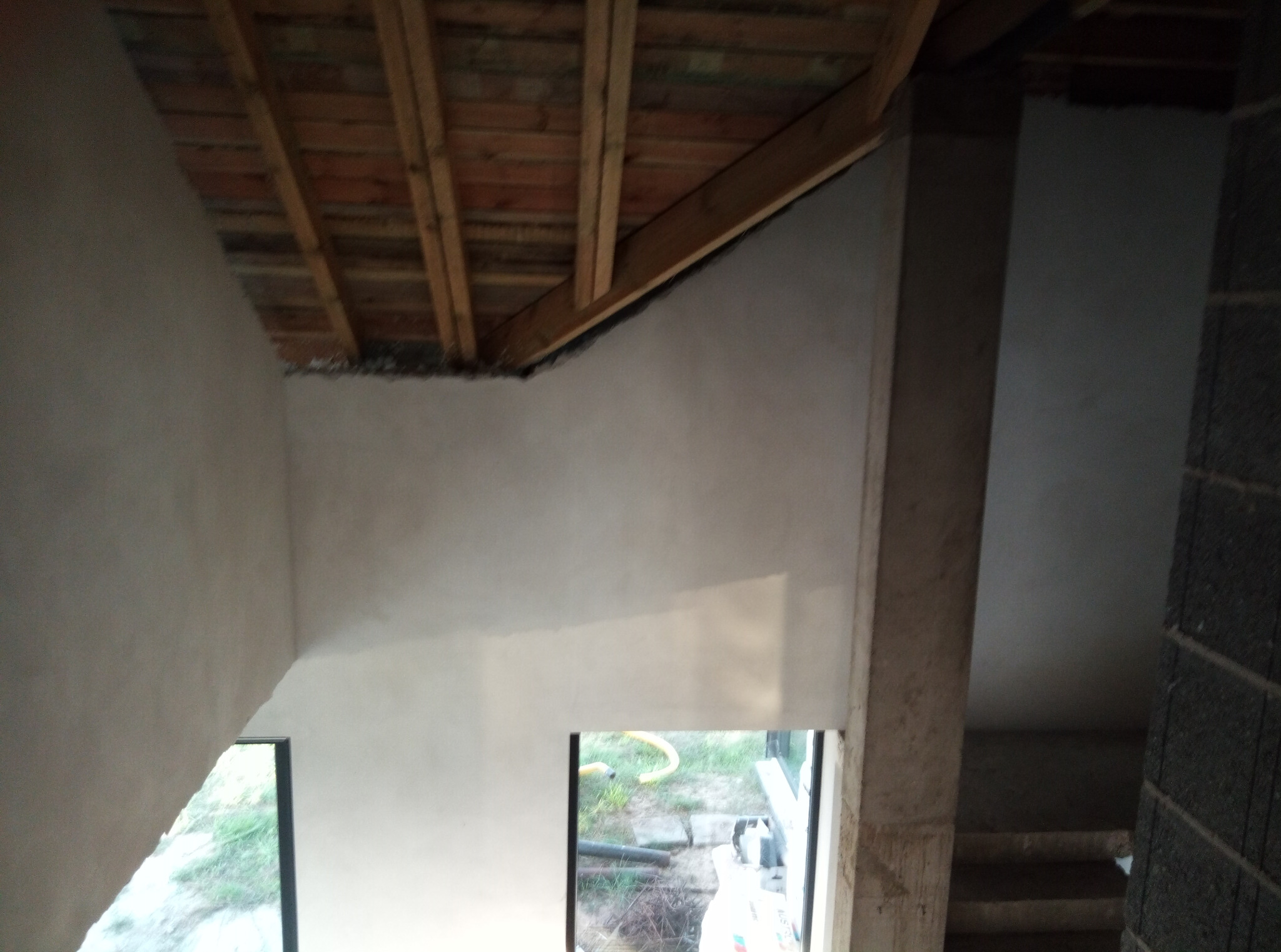 Surowe wnętrze z drewnianą konstrukcją dachu i otynkowanymi ścianami, widoczne okno z widokiem na zewnątrz. Etap prac wykończeniowych w domu.