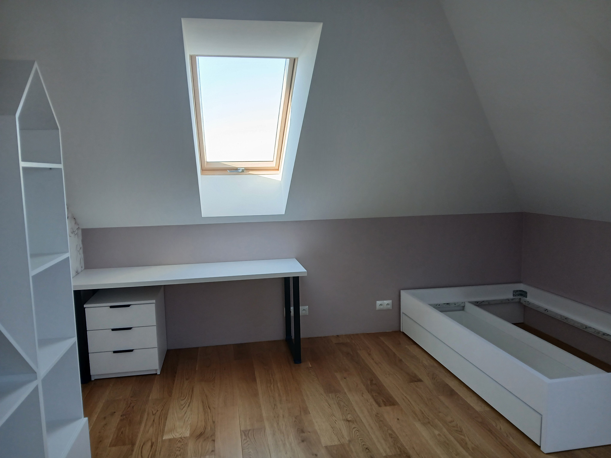 Pokój na poddaszu z oknem dachowym, białe meble: regał w kształcie domu, biurko z szufladami i rama łóżka. Ściany szare z bordowym pasem. Drewniana podłoga.