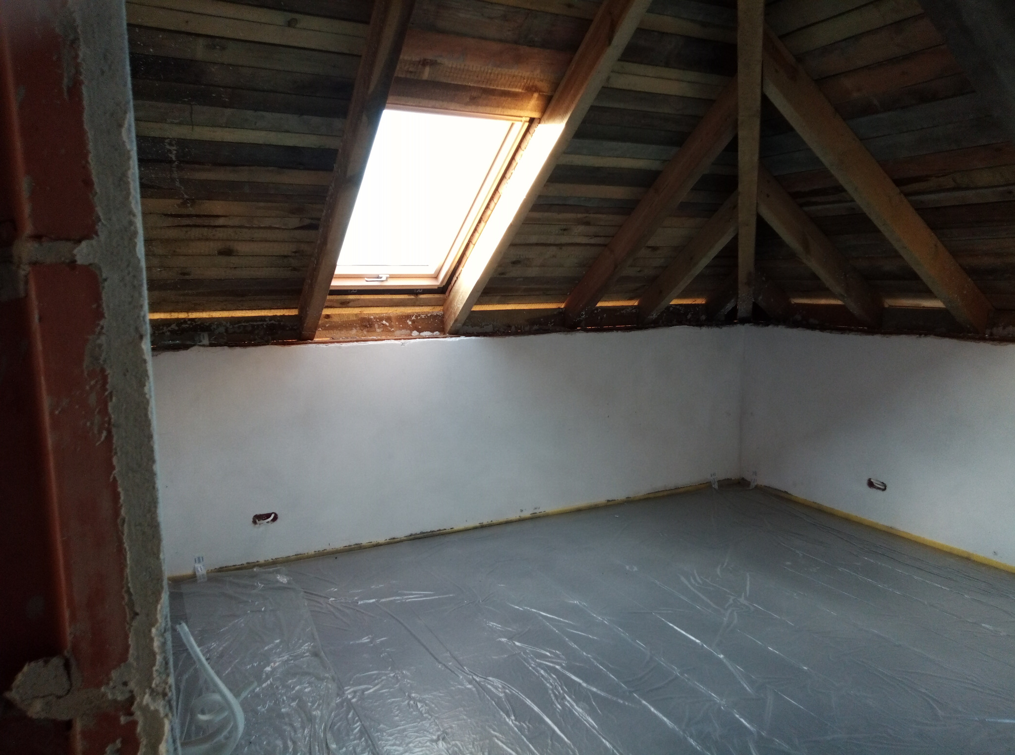 Poddasze w trakcie wykańczania: drewniana więźba dachowa z oknem połaciowym, otynkowane ściany, podłoga zabezpieczona folią malarską.