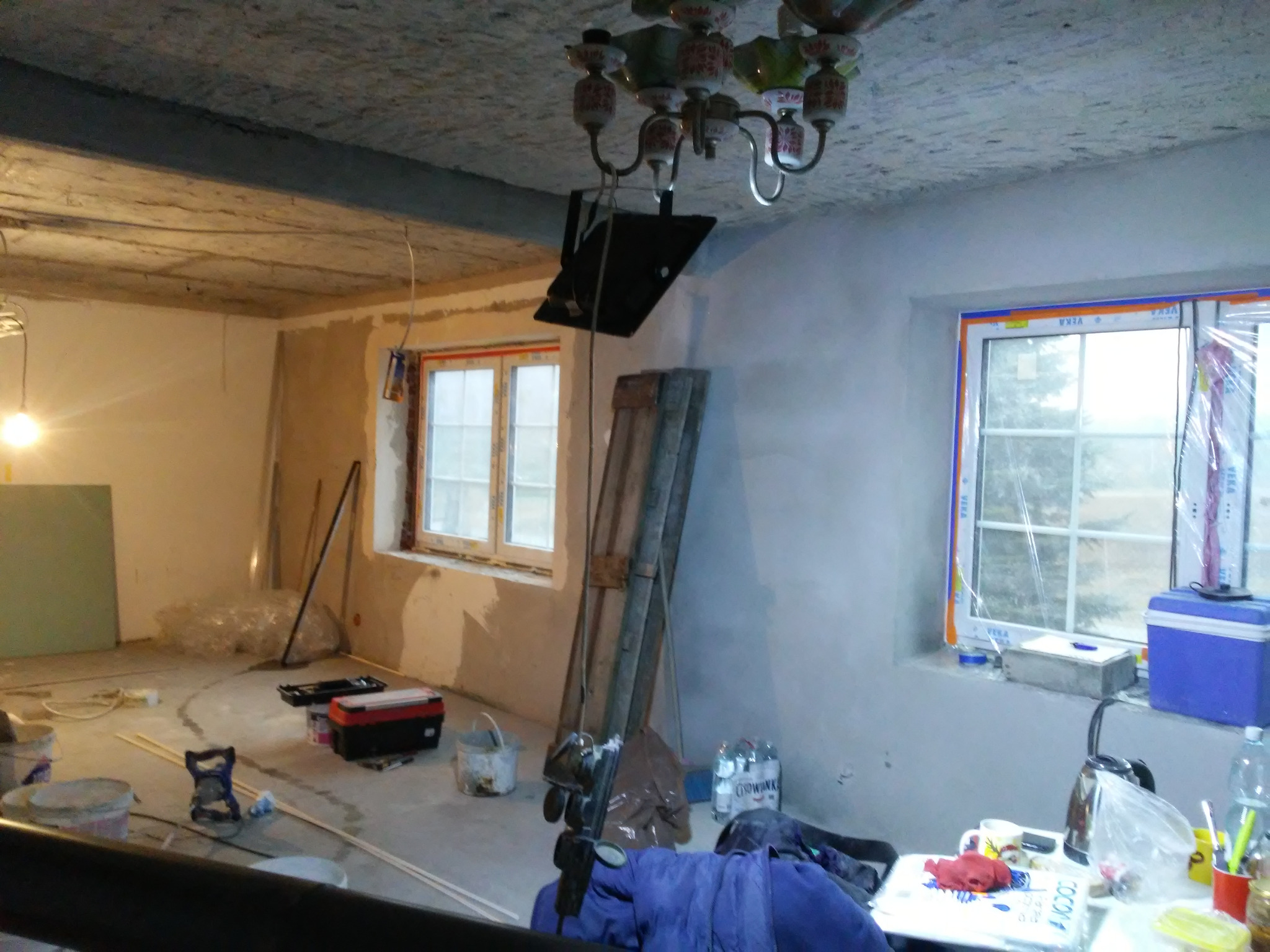 Wnętrze w trakcie remontu: surowy strop, ściany po szpachlowaniu, okna z folią ochronną, drabina oparta o ścianę, narzędzia i materiały budowlane.