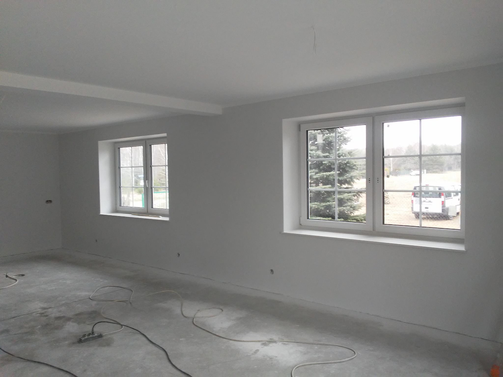Białe, świeżo pomalowane wnętrze z dwoma oknami i szarym betonowym podłożem. Widoczne kable i listwy przypodłogowe. Minimalistyczny styl, surowy stan wykończenia.