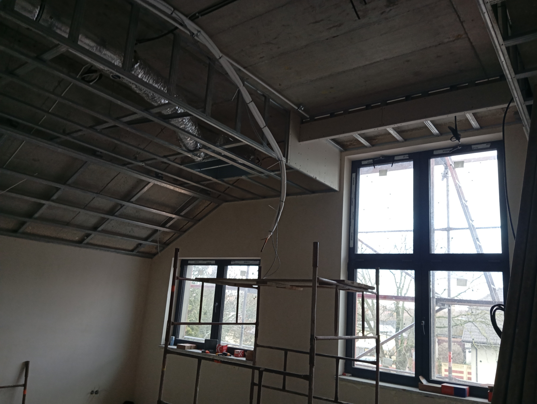 Stelaż sufitu podwieszanego z widoczną instalacją wentylacyjną i elektryczną w surowym wnętrzu. Dwa okna z widokiem na zewnątrz. Metalowe rusztowanie w pomieszczeniu.