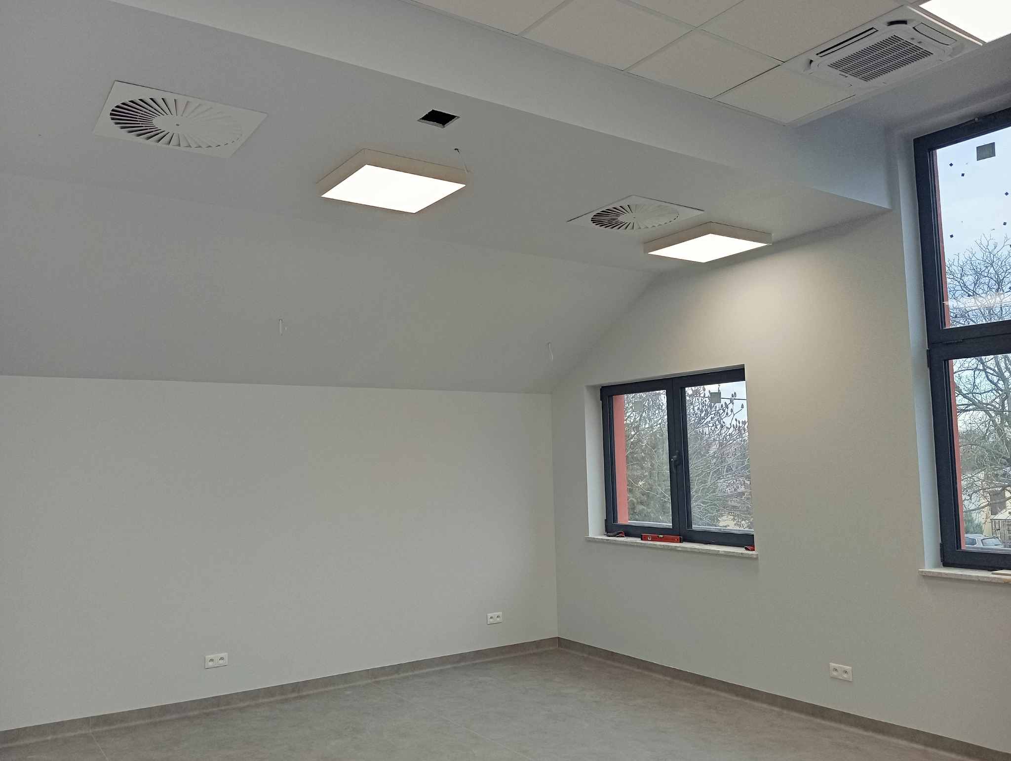 Jasne, świeżo pomalowane biuro z dwoma oknami, wentylacją sufitową i nowoczesnym oświetleniem. Widoczne gładkie ściany i szara podłoga.