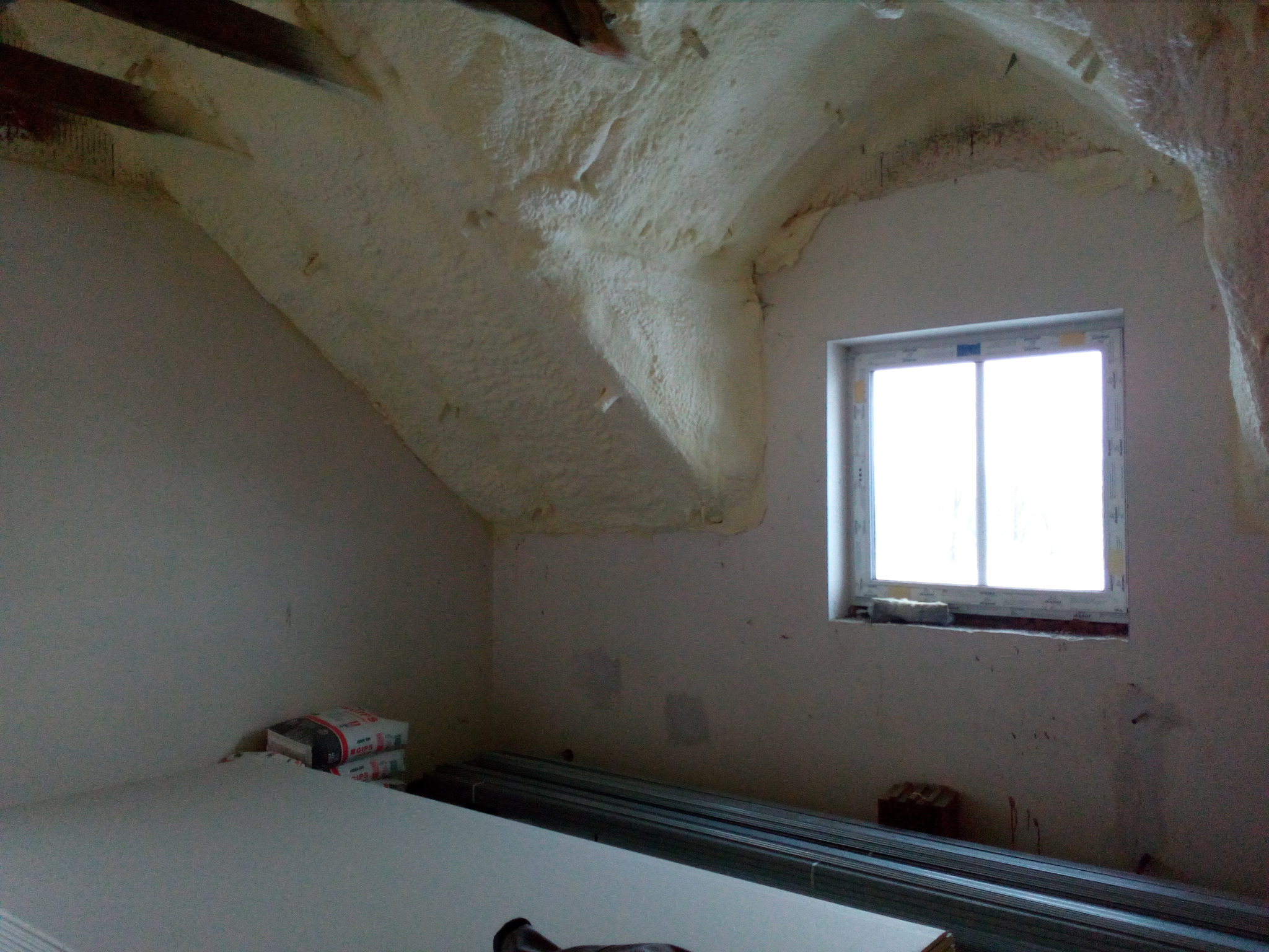 Poddasze po izolacji pianą PUR, widoczne płyty karton-gips i profile. Okno doświetla wnętrze. Etap prac wykończeniowych, przygotowanie do montażu płyt.