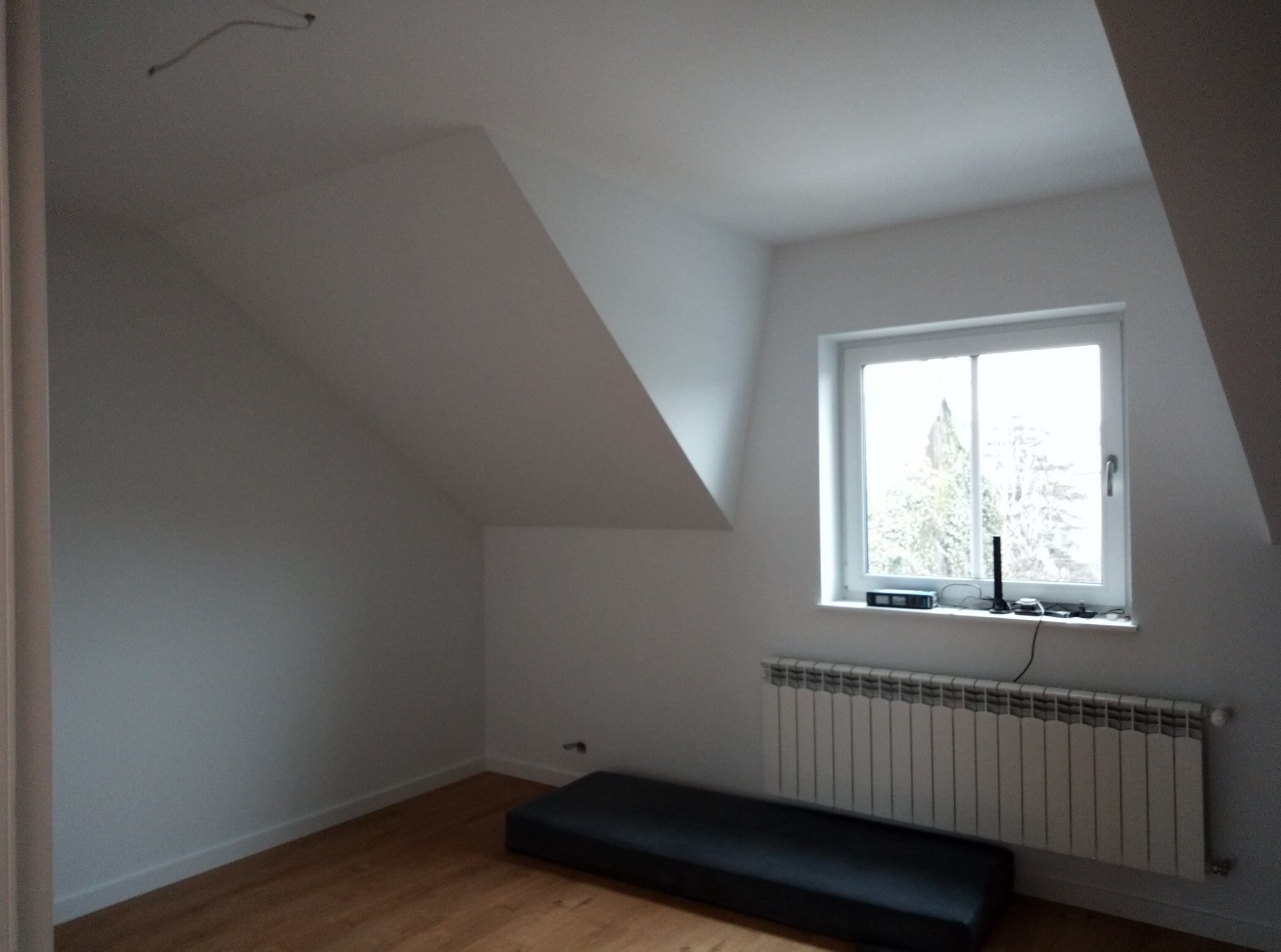 Poddasze po malowaniu na biało, z oknem, grzejnikiem i drewnianą podłogą. Widoczne skosy i gładkie ściany. Minimalistyczny styl wykończenia wnętrza.