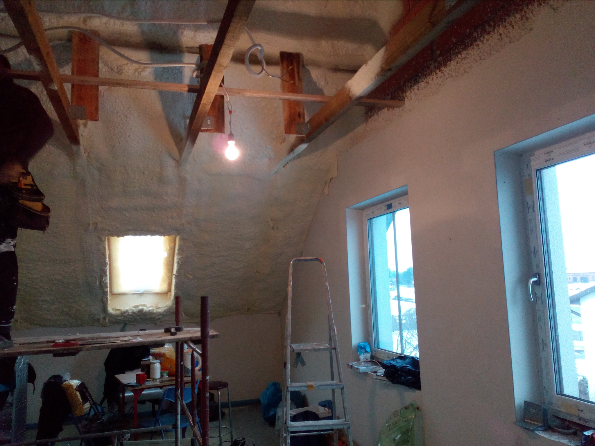Poddasze ocieplone pianą PUR, widoczne drewniane elementy konstrukcyjne, rusztowanie i drabina. Dwa okna w ścianie szczytowej doświetlają wnętrze. Prace wykończeniowe w toku.