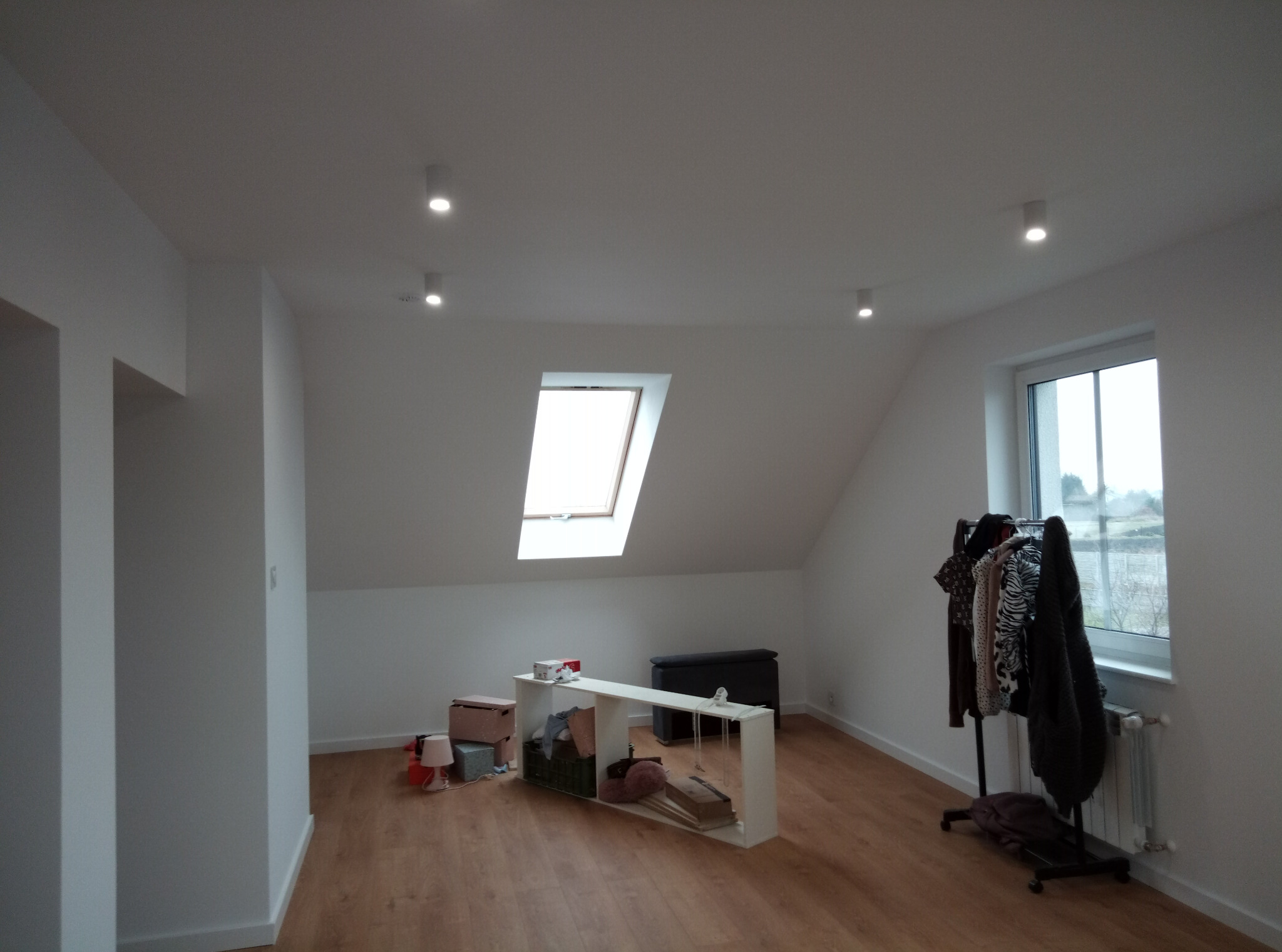 Jasne poddasze po remoncie: białe ściany, drewniana podłoga, okno dachowe i zwykłe okno. Wnętrze częściowo umeblowane, minimalistyczny styl.