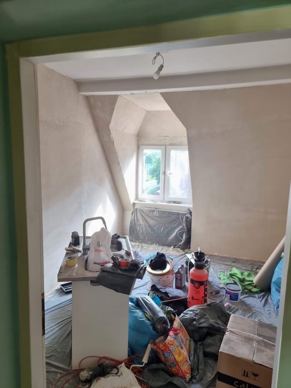 Pokój na poddaszu w trakcie prac wykończeniowych: ściany po gładzi, zabezpieczona podłoga, widoczny sprzęt malarski i narzędzia. Okno dachowe z widokiem na zieleń.