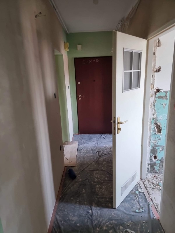 Korytarz w trakcie remontu: ściany częściowo otynkowane, zabezpieczona podłoga folią, otwarte drzwi z szybą. Widoczne ślady po starych instalacjach.