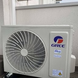 klimatyzator.net.pl - Zamontowana jednostka zewnętrzna klimatyzacji GREE na balkonie, widoczne czarne podkładki antywibracyjne. Biała ściana w tle, ujęcie z poziomu podłogi.