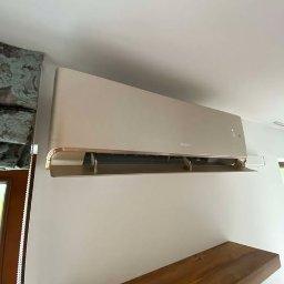 klimatyzator.net.pl - Beżowa klimatyzacja ścienna Gree w jasnym wnętrzu. Widoczna drewniana półka i fragment okna z ozdobną zasłoną. Minimalistyczny design, neutralne kolory.