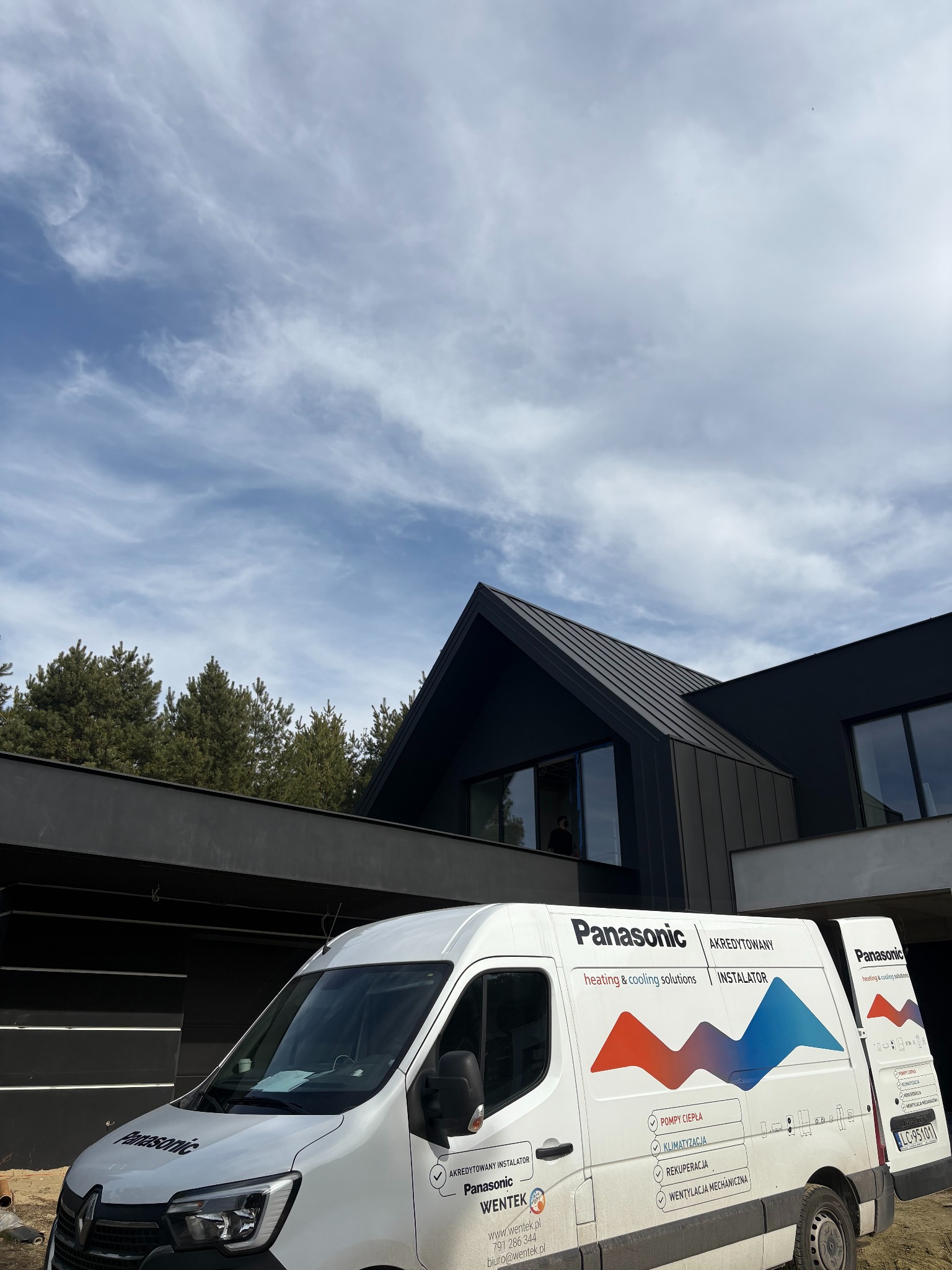 Biały van firmy Wentek z logo Panasonic na tle nowoczesnego, ciemnego domu z oknami. Usługi: pompy ciepła, klimatyzacja, rekuperacja, wentylacja mechaniczna.