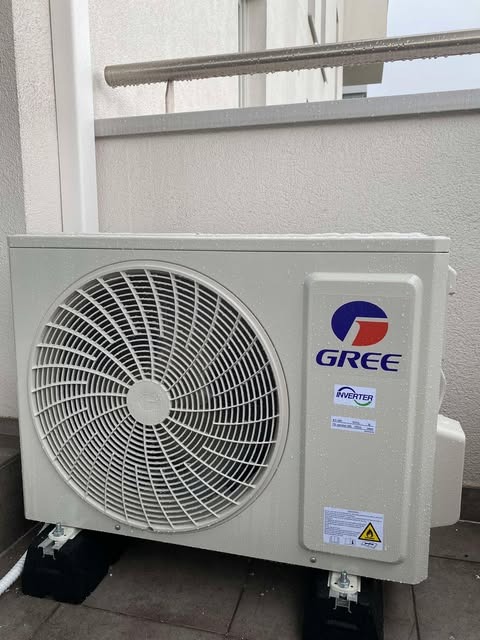 Zamontowana jednostka zewnętrzna klimatyzacji GREE na balkonie, widoczne czarne podkładki antywibracyjne. Biała ściana w tle, ujęcie z poziomu podłogi.