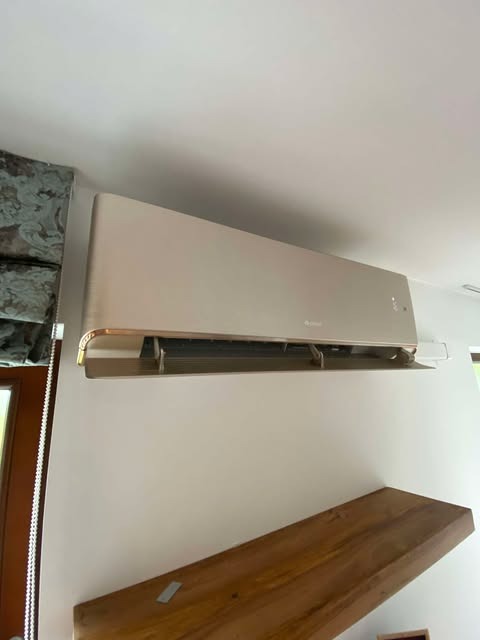 Beżowa klimatyzacja ścienna Gree w jasnym wnętrzu. Widoczna drewniana półka i fragment okna z ozdobną zasłoną. Minimalistyczny design, neutralne kolory.