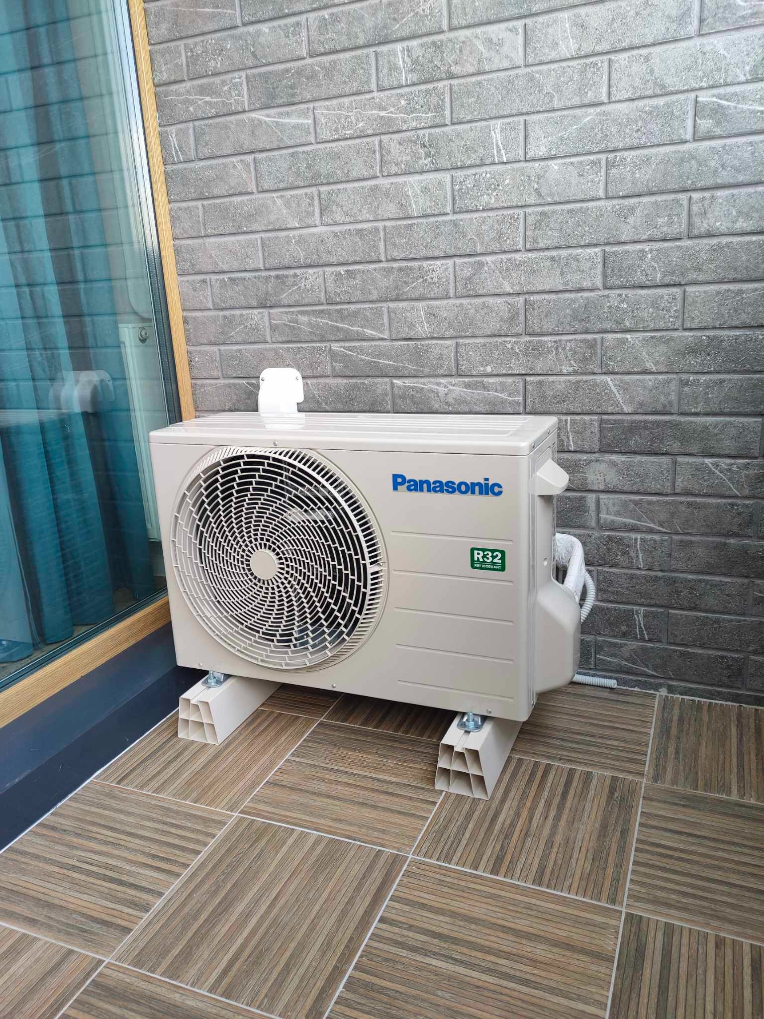 Zamontowana jednostka zewnętrzna klimatyzacji Panasonic R32 na tarasie, z widoczną elewacją z szarej cegły i podłogą z płytek imitujących drewno.