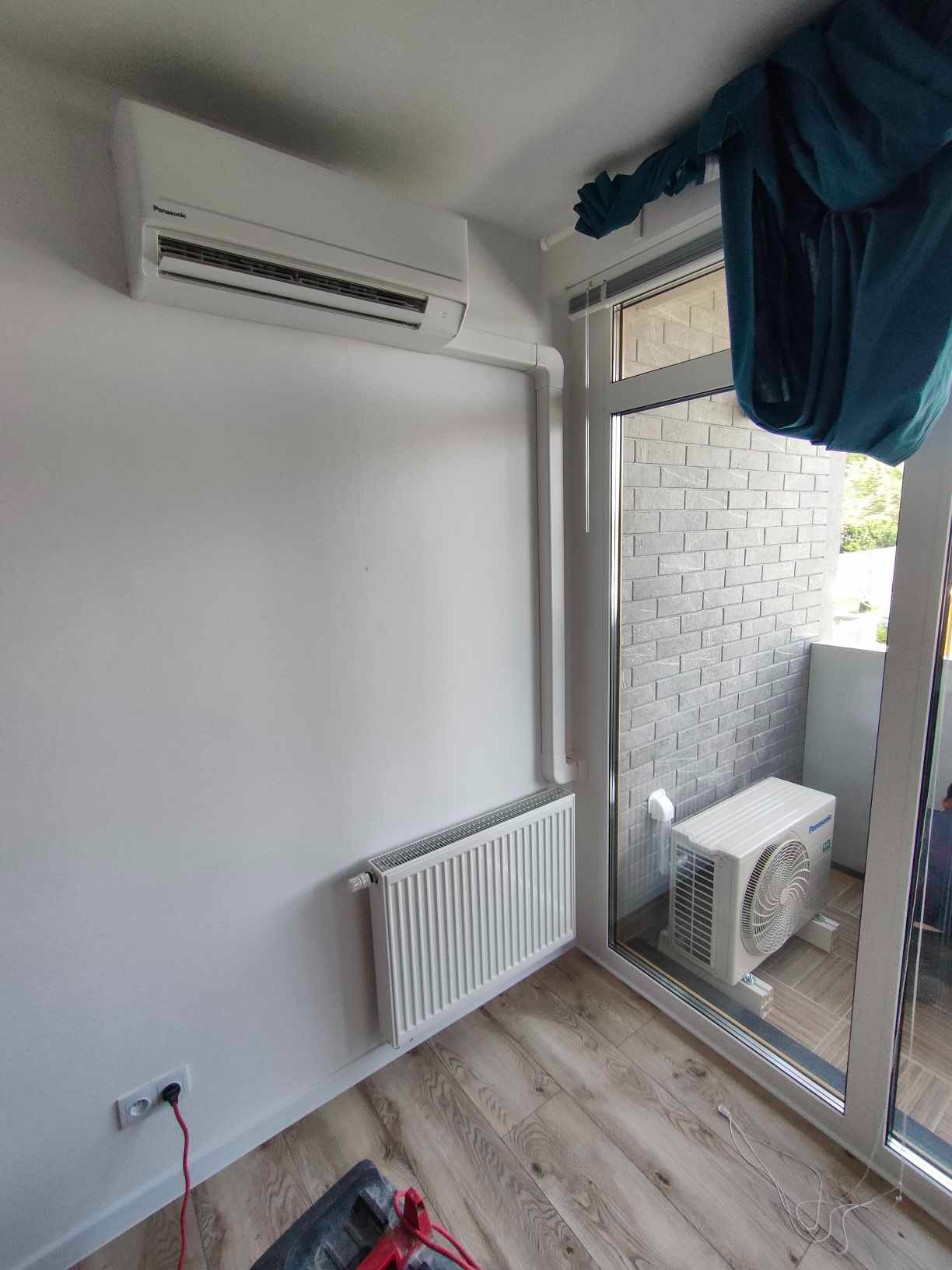 Biała klimatyzacja Panasonic zamontowana w jasnym pokoju z grzejnikiem i balkonem, widok z dołu, kabel zasilający widoczny przy podłodze.